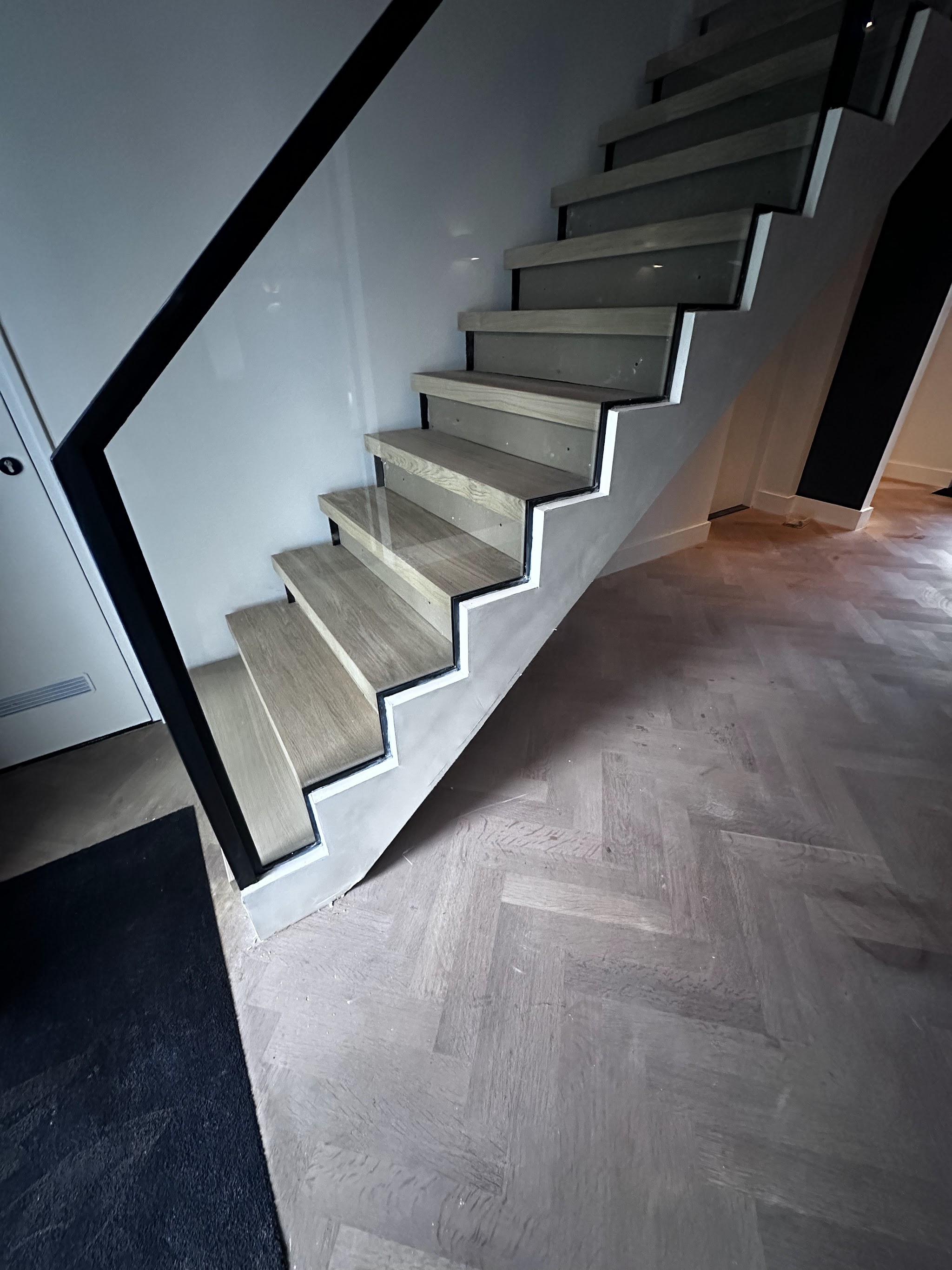 Een open trap met een glad gestucte boom, zwart stalen balustrade met glas, boven een visgraatvloer